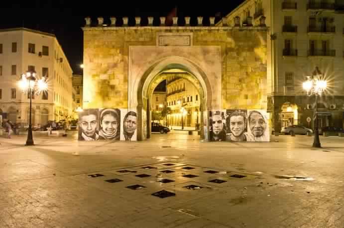 Retratos fotográficos de gran formato alineados frente a una puerta monumental iluminada de noche, proyecto INSIDE-OUT: Autocrazy in Tunisia.
